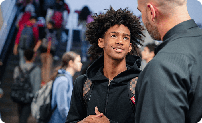A BARR student with a black hoodie and checkered backpack speaking to a teacher at school and smiling.