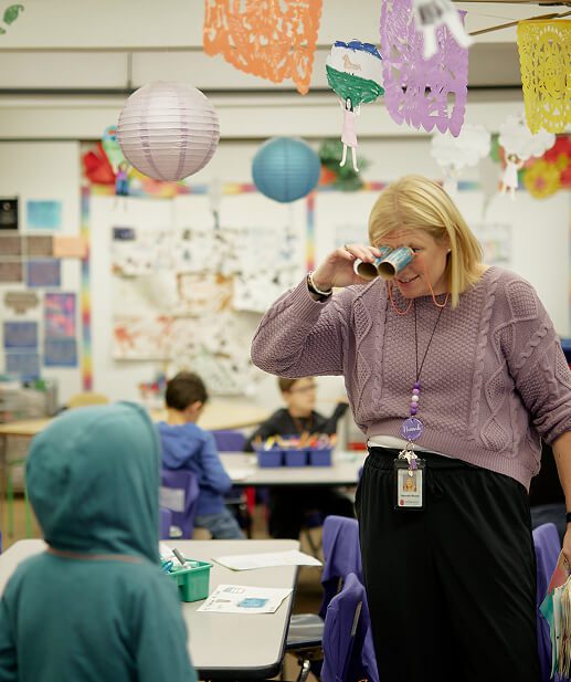 A BARR teacher looking at a student through binoculars made with arts and crafts materials.
