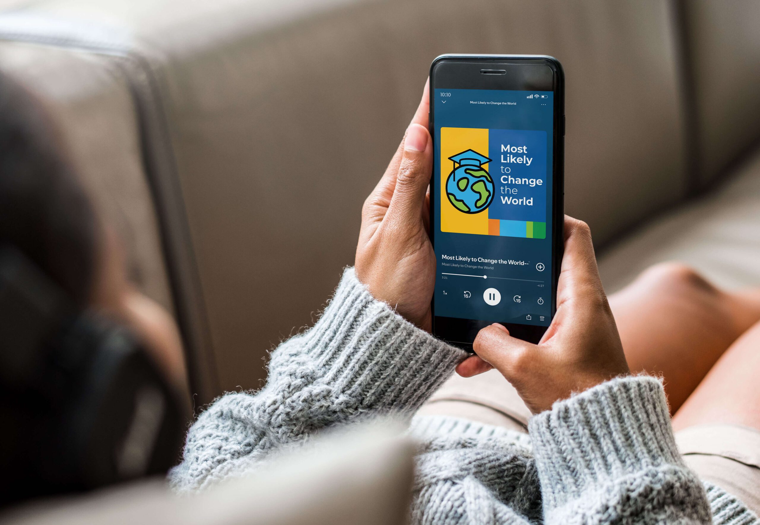 A person in a gray sweater scrolling on a cell phone listening to "Most Likely to Change the World" podcast